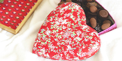 Herzförmige Schachtel mit rotem Blumenmuster von Décopatch, teilweise geöffnet, um die darin enthaltenen Pralinen zu enthüllen. Sie steht auf einer weißen Oberfläche neben einem Holztablett mit ähnlichem Blumenmuster.