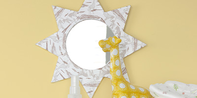 An einer gelben Wand hängt ein sternförmiger Spiegel von Décopatch mit weiß-braun gesprenkeltem Papier verziert. Davor steht eine dekorative gelbe Giraffenfigur mit weißen Flecken und einer Sprühflasche.