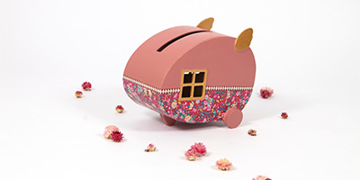 Eine rosa dekortierte Urne von Décopatch in Form eines Wohnwagens mit Blumenmuster und kleinen Rädern, geschmückt mit zwei ohrenähnlichen Vorsprüngen. Es ist von verstreuten rosa und weißen Blumen auf weißem Hintergrund umgeben.