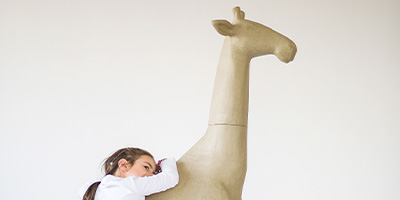 Ein junges Mädchen in einem weißen Hemd umarmt eine große, braune Giraffenskulptur von Décopatch vor einem schlichten weißen Hintergrund. Ihr Haar ist zurückgebunden und sie lehnt ihren Kopf an den Hals der Giraffe.