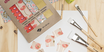 Ein Bastelset mit einem Paket gemustertem Décopatch-Papier mit floralen und geometrischen Mustern, mehreren Pinseln von Décopatch mit weißen Borsten und ausgeschnittenen Stücken Blumenpapier auf einer Holzoberfläche.