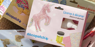 Décopatch-Set mit Einhorn-Dekoration. Die Box enthält eine weiße Einhornfigur aus Pappmaché, einen Behälter Klebtofflack und buntes Décopatch- Papier. Andere Sets mit unterschiedlichen Dekorationen sind teilweise im Hintergrund sichtbar.