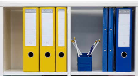 2 weiße Regalfächer in Nahaufnahme. Links stehen gelbe Ordner von Exacompta. Rechts ein Ordner, 2 Ringhefter und 1 mit Stiften gefüllter Stifteköcher in Blau von Exacompta.