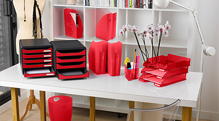 Homeoffice mit Schreibtisch und Regal in Weiß. Auf Schreibtisch stehen Schubladenboxen, Stehsammler, Stifteköcher und Briefablagen in Rot von Exacompta. Im Regal rote Stehsammler und Bücher. Außerdem stehen im Zimmer eine weiße Lampe, ein roter Papierkorb von Exacompta und ein transparenter Kunststoffstuhl.