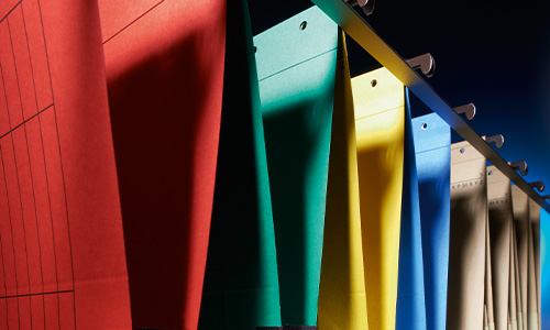 Bunte Hängeregistraturen von Falken in Rot, Grün, Blau, Gelb und Beige hängen in einer Reihe. Die Perspektive ist von einem Winkel aus, wodurch die Farbpalette und die Struktur der Hängeregistraturen hervorgehoben werden.