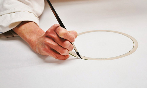 Eine Person zeichnet mit einem Pinsel einen Kreis auf weißes Papier. Ihre Hand ist ruhig und der Kreis ist teilweise vollständig. Sie trägt ein weißes Gewand und die Szene vermittelt Präzision und Konzentration. Es handelt sich um ein Hilfsmittel des Seidenmalspezialisten H Dupont.