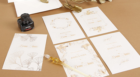 Eine Sammlung eleganter Hochzeitseinladungen mit Blumen- und Blattmotiven in Gold- und Beigetönen ist auf einer braunen Oberfläche ausgebreitet. Daneben liegen ein Tintenfass und ein dekorativer Goldstift mit transparenter Spitze. Von G. Lalo.