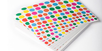 Ein Stapel weißer Stickerbogen, bedeckt mit bunten Punkt-Stickern in Rot, Gelb, Blau, Grün und Orange. Die Stickerbogen sind leicht aufgefächert auf einer weißen Oberfläche.