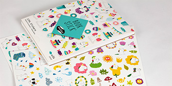 Bunte Aufkleber mit verschiedenen skurrilen Motiven wie Tieren, Früchten und anderen verspielten Elementen sind ausgebreitet. In der Nähe liegt ein Heft mit der Aufschrift „Stick Book“, das teilweise geöffnet ist und in dem noch weitere Aufkleber zu sehen sind.