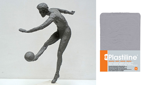 Neben einer Packung Plastiline-Modelliermasse ist eine graue Skulptur einer Person zu sehen, die einen Ball kickt. Die Skulptur stellt eine dynamische Bewegung dar, wobei die Figur auf einem Bein balanciert und das andere Bein nach vorne streckt, um den Ball zu kicken. Sie wurde aus der hochpräzisen Modelliermasse Plastiline hergestellt.