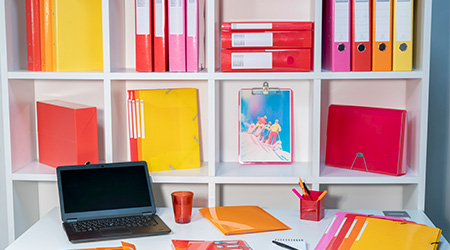 Weißes Regal mit Ordnern, Archivboxen, Sammelmappen, Klemmbrett und Fächermappe von Exacompta in Rosa, Rot, Orange und Gelb. Davor ein weißer Schreibtisch mit Sammelmappen, Ordungsmappe, Sichtmappe, Stifteköcher von Exacompta und schwarzem Laptop.