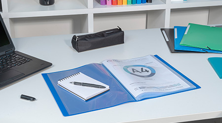 Ein Schreibtisch mit einem Laptop, einem schwarzen Federmäppchen, einem blauen Ordner mit einem A4-Blatt und einem Notizbuch darin, einem Stift und mehreren bunten Ordnern im Hintergrund. Die Produkte sind von Exacompta.