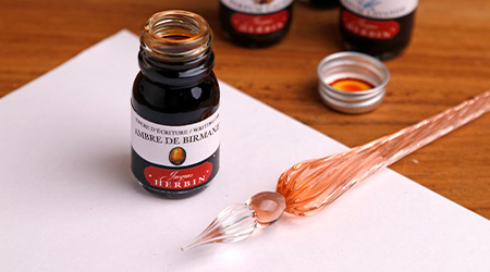 Offener 10ml-Flakon der Tinte Ambre de birmanie und Glasfeder von Jacques Herbin auf weißem Blatt. Alles liegt auf Holztisch.