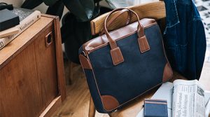 Braun-blaue, mittelgroße Ledertasche mit Stoffelementen und Henkel von Quo Vadis auf Holzstuhl in Zimmer einer belebten Wohnung. Jacke hängt am Stuhl.