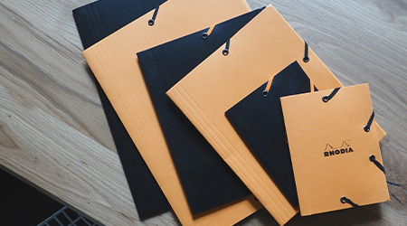 Eckspannmappen von Rhodia in schwarz und orange in verschiedenen Formaten versetzt übereinandergestapelt. Auf braunem Holztisch. Blick von oben.