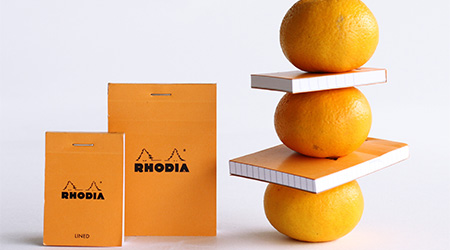 2 orangene, stehende Blöcke von Rhodia. Rechts daneben 3 übereinander gestapelte Orangen, zwischen denen 2 orangene Rhodia-Blöcke stecken. Tisch und Fläche sind weiß.