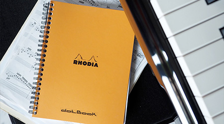 Oranges Spiralbuch dotBook von Rhodia auf Stuhl auf Notenblättern. Rechts oben Klaviertastatur.