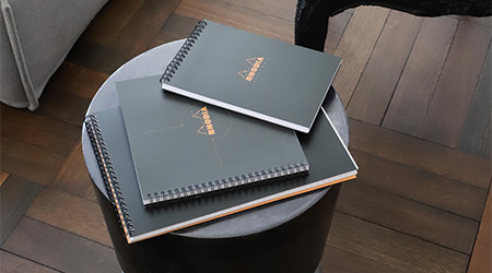 3 unterschiedlich große, schwarze Spiralbücher von Rhodia auf schwarzem Hocker in Zimmer mit Holzboden.