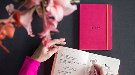 Frau schreibt an schwarzem Tisch in Kalender Perpetual von Rhodia. Davor liegt geschlossenes Kalenderbuch in pink. Man sieht nur die Hände. Blick von oben.