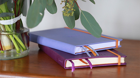 Notizbücher in 2 verschiedenen Violett-Farbtönen auf dunkelbraunem Holzschrank. Links Blumen in einer Vase.