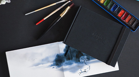 Schwarzes Skizzenbuch Rhodia Touch mit Aquarellfarben und Pinsel auf schwarzem Hintergrund. Dazu offenes Skizzenbuch mit Aquarellbild.