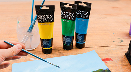 Tuben Acrylfarbe von Blockx in Gelb, Grün und Blau auf Holztisch. Hand hält Pinsel in der Hand und malt Landschaftsbild.