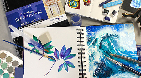 Tisch mit Skizzenbuch Mixed Media Beta von Stillman&Birn, schwarzer Spiraleinband. Mit Stiften, Pinseln und Farbe. Offenes Skizzenbuch mit blauen Zeichnung von Wellen und Blumen.