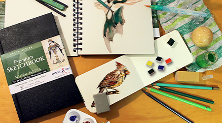 Tisch mit Skizzenbuch Mixed Media Delta von Stillman&Birn, schwarzer Einband. Mit Pastellkreide und Farbe. Offene Skizzenbücher mit Zeichnung eines Vogels und einer Tänzerin.
