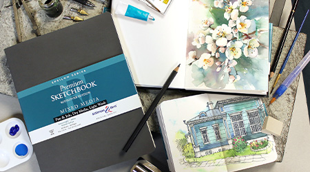 Tisch mit Skizzenbuch Mixed Media Epsilon von Stillman&Birn, grauer Einband. Mit Stiften. Offene Skizzenbücher mit Zeichnung von Blumen und eines Hauses im Grünen.