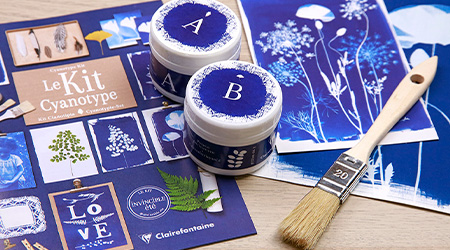 Blaues Cyanotypie-Set auf Holztisch. 2 Blatt Papier, die mit blauem Muster bedruckt sind, 1 Heft, 1 Borstenpinsel, 1 Dose Ammoniumeisencitrat und 1 Dose Kaliumferricyanid.