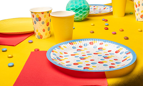 Partytisch mit gepunkteten Papptellern und -bechern, gelben Bechern und roten Servietten auf gelber Tischdecke.