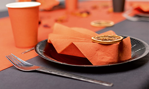 Auf einem Teller gefaltete orangefarbene Serviette mit einer getrockneten Orangenscheibe, daneben eine Plastikgabel und ein Pappbecher.
