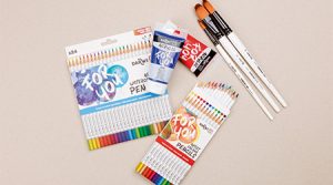 Malutensilien wie Buntstifte, Pinsel und eine Tube Acrylfarbe auf einer beigen Fläche.